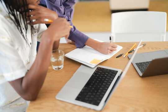 Two people work together at a modern office desk. One writes notes in a notebook, while the other person listens intently. Laptops and stationery are spread across the wooden surface.