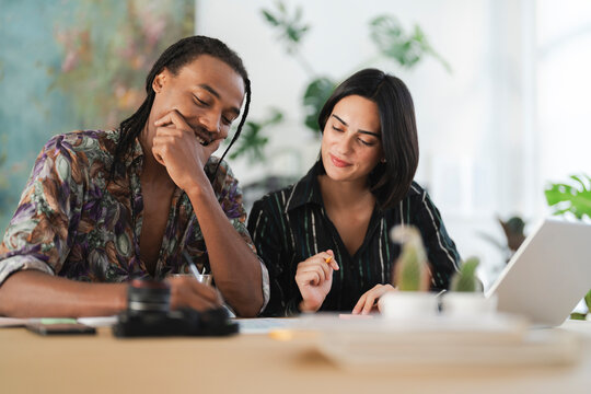 A diverse couple or colleagues are collaborating at a table in a bright office. The man smiles while the woman holds a pencil, focusing on documents. They are planning or working together.