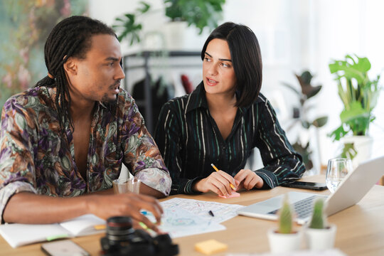 Two diverse colleagues collaborate at a modern office desk. They are discussing project plans, with a laptop, camera, and documents spread out. They are focused on their work.