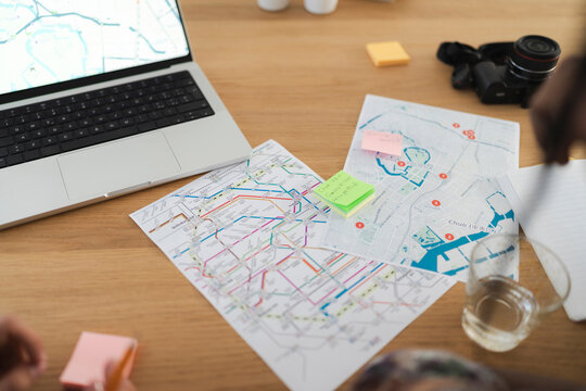 People plan a trip using a laptop, physical maps, and sticky notes on a wooden table. A camera and notebook are also present, indicating travel preparation.