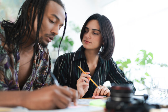 Two diverse young professionals collaborate on a project at a desk. The man writes while the woman observes, holding a pencil. They are focused on their work.