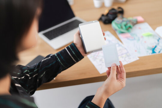 A person holds a portable instant photo printer and a blank photo print. A laptop and papers are on a wooden desk, suggesting a creative or memory-making activity.