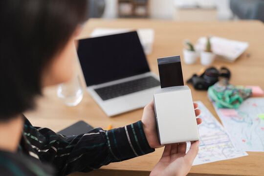 A person holds a compact photo printer, watching as a freshly printed picture emerges from the device. A laptop and travel maps are visible on the wooden desk.