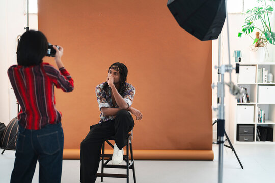 A photographer captures a male model posing on a stool in a professional photo studio. The model wears a patterned shirt and headband, looking thoughtfully away.