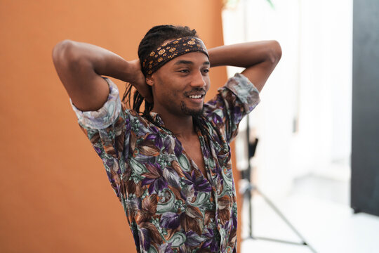 Young Black man with dreadlocks and a patterned headband smiles while posing with his hands behind his head in a studio setting. He wears a colorful shirt.