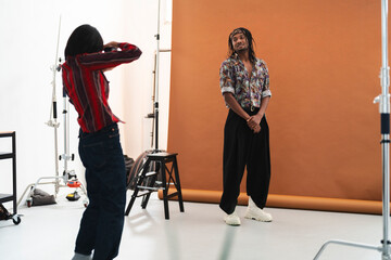 A photographer captures a smiling male model during a studio photoshoot. The model poses against a brown backdrop with professional lighting equipment around.