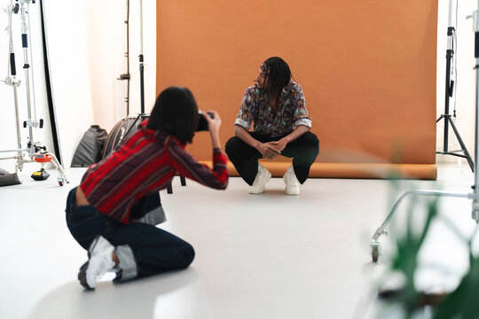 A female photographer kneels, taking photos of a male model squatting against a brown backdrop in a professional studio. The model poses for the camera.