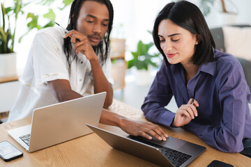 Two diverse colleagues collaborate on laptops in a bright office. They are focused on their work, sharing ideas and discussing project details. Professional teamwork.