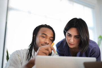 Two diverse colleagues, a man and a woman, collaborate closely, intently focused on a laptop screen. The man points with a pen, sharing insights with his attentive female coworker.