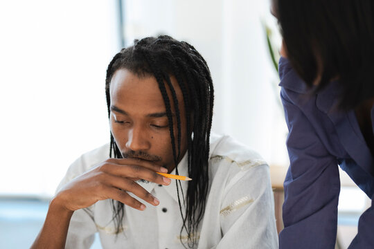 A focused man with dreadlocks holds a pencil to his chin, deep in thought in a bright office. Another person stands nearby, observing. This depicts concentration and teamwork.