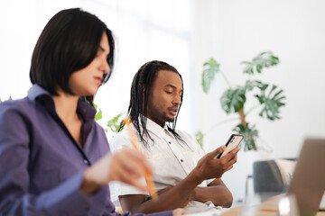 Two professionals work in a bright office setting. A man with dreadlocks checks his smartphone, while a woman in the foreground holds a pencil, engaged in her work.