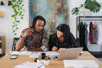 Two diverse professionals collaborate on a creative project at a modern workspace. They review documents and maps, utilizing a laptop for their focused work. Productive team.