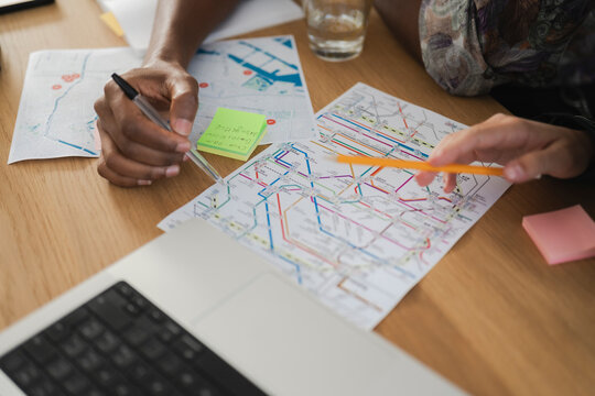 Two individuals are planning a route or trip, examining maps and notes on a wooden desk. One points with a pen, the other with a pencil, collaborating on their journey.