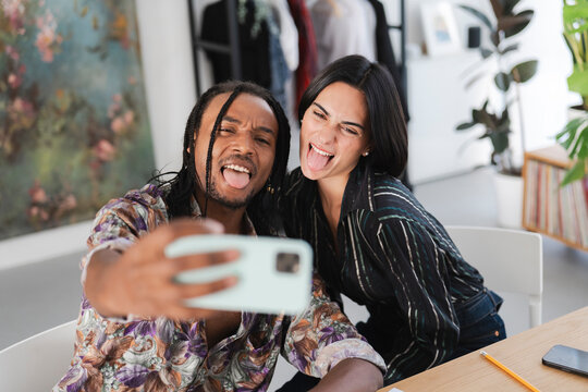 A diverse young man and woman take a playful selfie together, sticking out their tongues and making funny faces. They are enjoying a fun, candid moment indoors.