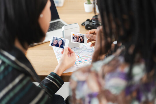 Two diverse friends review instant photos they just printed from a portable device. They are sitting at a desk with a laptop and camera, planning their next adventure using a map.