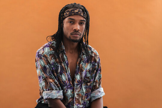 A young Black man with dreadlocks and a patterned bandana looks directly at the camera with a calm expression. He wears a colorful, unbuttoned shirt against a plain orange background.