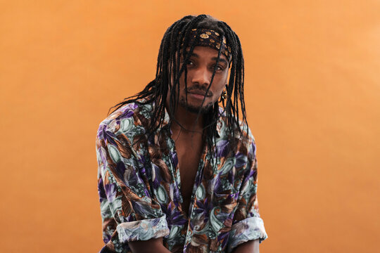 A young Black man with dreadlocks and a patterned bandana gazes directly at the viewer. He wears a vibrant, open shirt against a plain orange background. He has a beard and an earring.