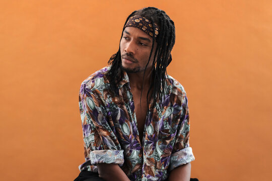Stylish young man with dreadlocks and a patterned bandana gazes thoughtfully to the side. He wears a vibrant patterned shirt against a warm orange background.