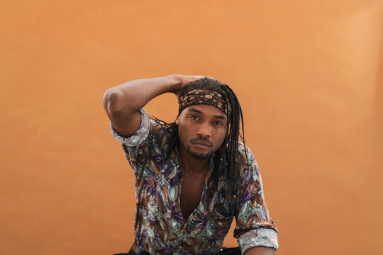 A young Black man with dreadlocks and a patterned headband poses against an orange background. He looks directly at the camera with a serious expression, hand on his head.