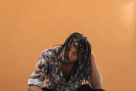 A young Black man with dreadlocks and a bandana looks directly at the camera. He wears a colorful patterned shirt, posing thoughtfully against a simple orange background.
