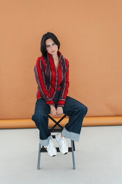 A stylish young woman with dark hair sits confidently on a stool, gazing directly at the camera. She wears a striped shirt, wide-leg jeans, and white athletic shoes in a studio.