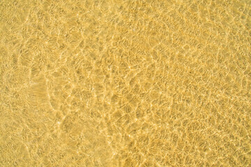 A background of clear water over a sandy bottom. Shallow water with yellow sand. Gentle ripples on the surface. Glittering reflections on the sea.