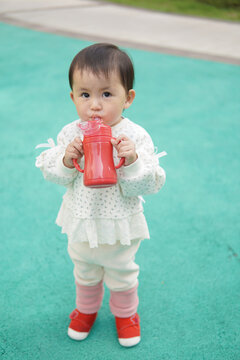 A toddler in a white dotted outfit holds a red sippy cup on a green playground surface
