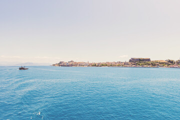 Fototapeta premium Corfu sunlit shoreline and deep blue sea frame docked ships and distant buildings, Greece