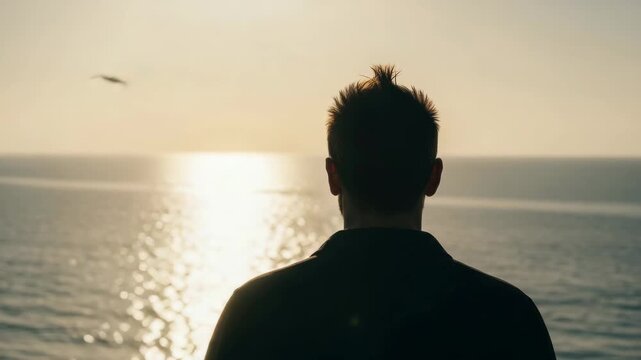 Silhouette of a young adult male from behind watching the sunset over the ocean. Contemplative person with a mohawk hairstyle enjoying a peaceful moment of solitude and freedom