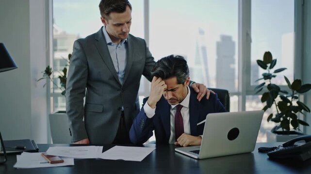 Colleague comforting stressed businessman in modern office at daytime, support and leadership empathy during financial setback and deadline pressure with paperwork and laptop on desk