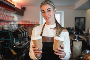Blonde waitress with a warm smile presents a to go coffee as the cafe owner prepares drinks behind the counter in a busy shop
