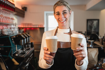 Friendly blonde waitress offers a takeaway coffee while the cafe owner stands behind the counter in a bright business atmosphere