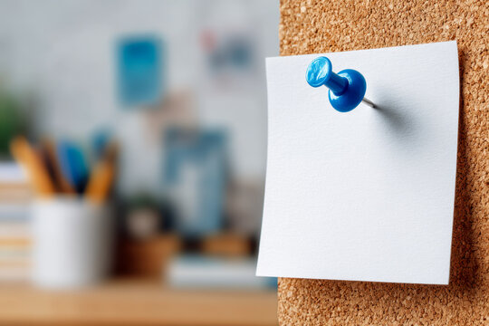 Blank white note paper pinned with a blue pushpin on a corkboard in a cozy and blurred home office setting with stationery in the background