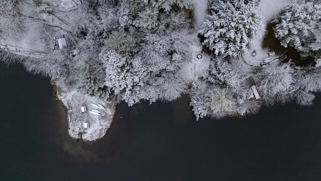 Aerial view of snow-laden trees and canoes: a tranquil winter scene where the forest meets the dark, still water's edge, forming abstract patterns. White Mountains, NH, USA.