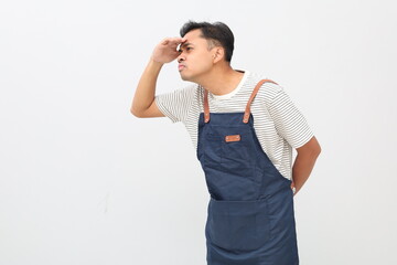 Fototapeta premium Portrait of a curious Asian man in an apron, holding his hands over his eyes and peering into the distance, looking far away, expecting and searching for someone, isolated on a white background.