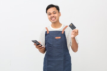 Successful young Asian man, barista, bartender, coffee shop employee wearing blue apron, holding mobile phone, credit card and bank card, isolated on white background, smiling looking at camera.