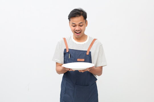 Surprised expression of young Asian male chef or waiter wearing apron with empty plate on white background, cooking or culinary concept. - Powered by Adobe