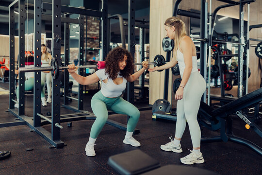 Two women are training together in a gym, with one performing a barbell squat while the other acts as a spotter