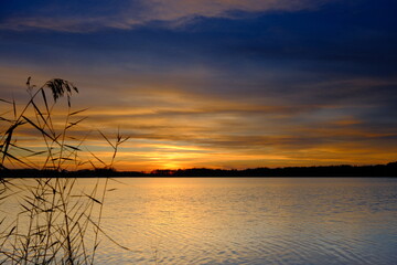 Zachód słońca nad spokojnym jeziorem, sfotografowany bez długiego naświetlania. Widoczna struktura fal, dynamiczne trzciny na pierwszym planie oraz intensywne kolory nieba tworzą naturalną, reportażow © Przemysaw