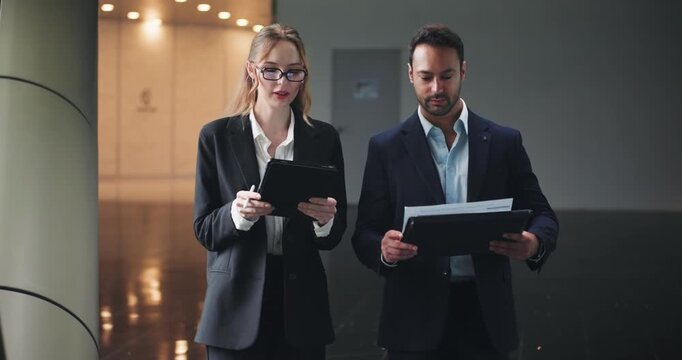 European Businessman Listening To Businesswoman With Tablet Discussing Report While Walking In The Office Corridor. - wide shot