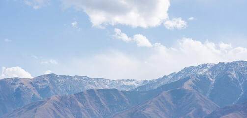 mountainous terrain under a clear blue sky. The rolling foothills, cloaked in auburn hues, contrast with the rugged, snow-dusted peaks in the distance. scattered clouds adorning the sky