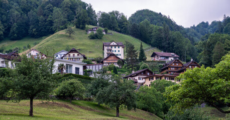 Houses scattered across a green hillside in a mountainous alpine village, surrounded by dense forest and meadows. Traditional chalets blend into the natural landscape under a cloudy sky.