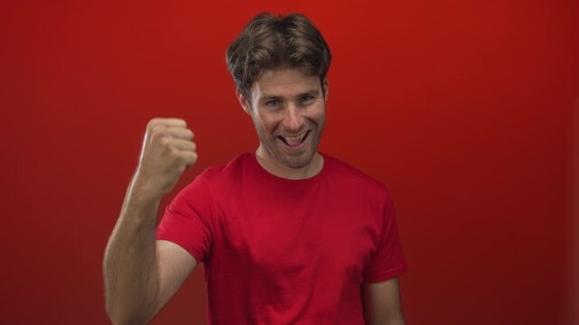 Young man in red t shirt smiling and raising clenched fist in studio with red backdrop; excitement confidence success.