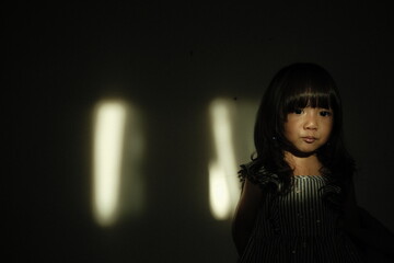 Portrait of a young girl standing against a dark background with cinematic light streaks on the wall. She makes a pout expression, creating a dramatic and artistic mood.