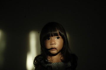 A young girl stands in a dark room with dramatic window light illuminating her face. Her eyes look upward with a subtle anxious expression, while shadows create a cinematic and emotional atmosphere.