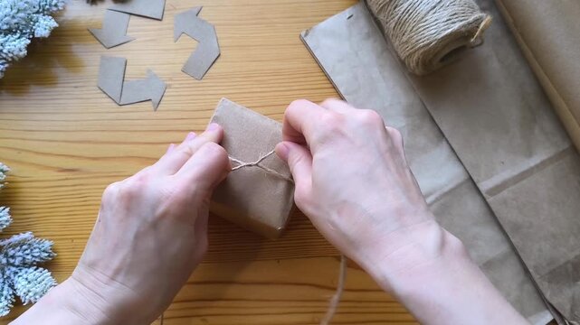 Gift wrapping in eco-friendly materials against the background of a recycling symbol and Christmas tree branches. Tie the gift with twine.