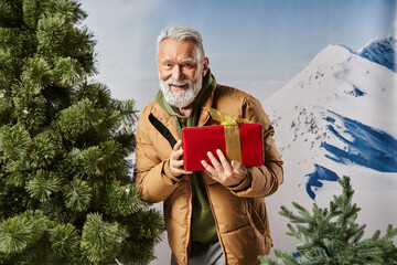 Joyful senior man holding a gift in a winter wonderland setting with snow capped mountains