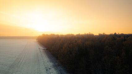 Atmospheric aerial view at sunrise with warm golden light illuminating an autumn forest and a river in the background. The morning fog and the glowing horizon create a calm, peaceful mood in this scen