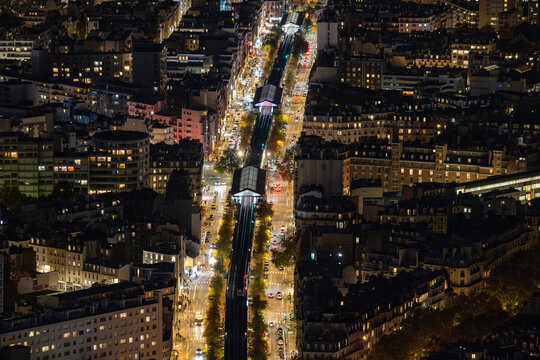 Aerial view of the brightly lit elevated railway slicing through the urban fabric, flanked by buildings, trees, and streets illuminated at night, Paris, Ile-de-France, France.