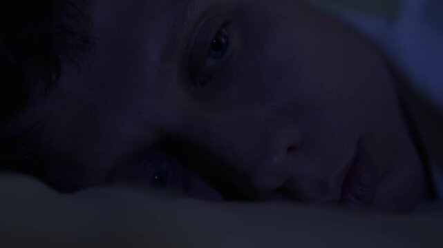 A lonely, sleepless man lies awake at night and stares into the dark. Feeling emotional emptiness, sadness, and life disappointment. Dark moody bedroom.